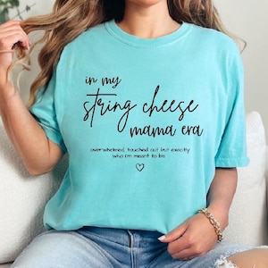 May include: A light blue t-shirt with the text "in my string cheese mama era" in black script. Below the text is the phrase "overwhelmed, touched out but exactly who I'm meant to be" and a small heart.