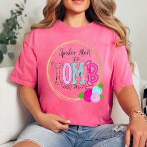 May include: A pink t-shirt with the text "Spoiler Alert the Tomb was empty" in a gold circle. The word "TOMB" is in teal and pink letters. Floral accents in pink, blue, and green are at the bottom.