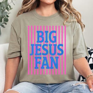 K&ouml;nnte beinhalten: Olivgr&uuml;nes T-Shirt mit dem blauen Schriftzug "BIG JESUS FAN" vor einem Hintergrund aus pinkfarbenen vertikalen Streifen. Das T-Shirt hat einen l&auml;ssigen Stil.