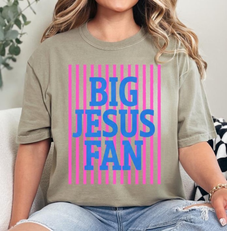 K&ouml;nnte beinhalten: Olivgr&uuml;nes T-Shirt mit dem blauen Schriftzug "BIG JESUS FAN" vor einem Hintergrund aus pinkfarbenen vertikalen Streifen. Das T-Shirt hat einen l&auml;ssigen Stil.