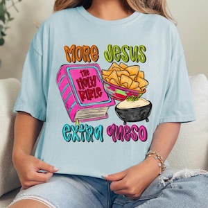 May include: Light blue t-shirt with the text "MORE JESUS EXTRA QUESO". The graphic includes a pink Bible, a bowl of tortilla chips, and a bowl of queso. The text and graphics are in bright colors.