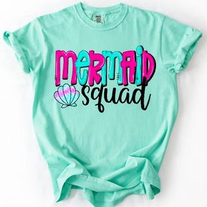 May include: A mint green short-sleeve t-shirt featuring the text "MERMAID squad" in a bold, layered font. The word "MERMAID" is in bright pink and turquoise, while "squad" is in black script. A small, colorful seashell icon is included.