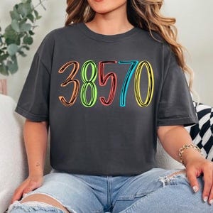 May include: Dark gray t-shirt with the number "38570" in colorful, outlined lettering. The shirt is worn with blue jeans. The numbers are outlined in orange, green, red, blue, and yellow.