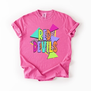 May include: A bright pink short-sleeve t-shirt featuring the text RED DEVILS in colorful block letters. The design includes geometric shapes in teal, magenta, purple, and lime green, layered over a black and white patterned background.