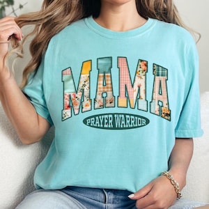 May include: A light blue t-shirt with the word "MAMA" in a patchwork design, featuring floral and plaid patterns. Below the word "MAMA" is the text "PRAYER WARRIOR" in a green oval. The shirt is a comfortable, casual style.