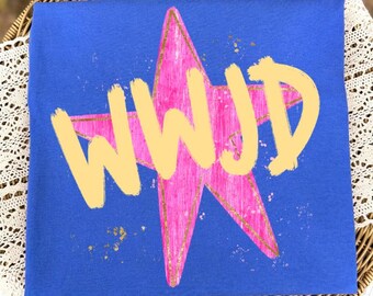 Puede incluir: Una camiseta azul real que presenta un gráfico de una estrella rosa grande y texturizada con contornos y motas de color dorado. Las letras WWJD están impresas en el centro de la estrella en una fuente negrita de color amarillo claro estilo pincel.
