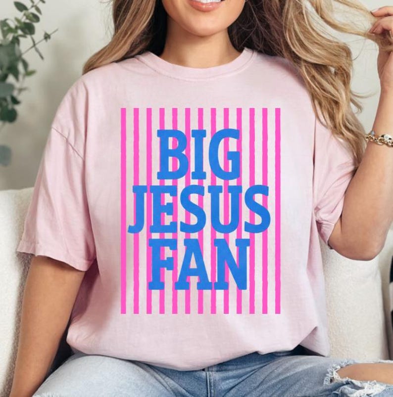 K&ouml;nnte beinhalten: Hellrosa T-Shirt mit dem Schriftzug "BIG JESUS FAN" in blauen Blockbuchstaben. Der Text befindet sich vor einem Hintergrund aus vertikalen rosa und wei&szlig;en Streifen. Das T-Shirt hat kurze &Auml;rmel.