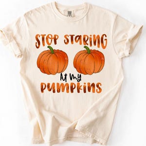 May include: A cream-colored short-sleeve t-shirt featuring two orange pumpkins centered on the chest. The text "STOP STARING" is printed in orange above the pumpkins, and the text "AT MY PUMPKINS" is printed in black and orange below them.