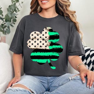 May include: Dark gray t-shirt featuring a large shamrock design. The shamrock is divided into sections with an American flag pattern and green and black stripes. The shirt is a casual style, suitable for St. Patrick's Day.