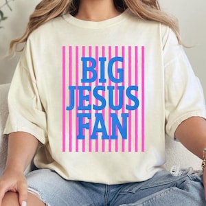 K&ouml;nnte beinhalten: Cremefarbenes T-Shirt mit dem Text "BIG JESUS FAN" in blauen Blockbuchstaben, vor einem rosa gestreiften Hintergrund. Das Shirt hat eine lockere Passform und kurze &Auml;rmel.