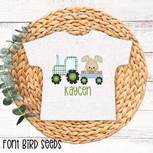 May include: A light gray t-shirt featuring a cartoon tractor with a plaid design and a bunny in a wagon. The tractor has green wheels and the name "kaycen" in green script below the image. The shirt is displayed on a woven basket.