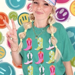 May include: A green t-shirt featuring a grid of nine colorful cowboy boots in shades of pink, yellow, and green. The shirt is paired with a pink and white striped baseball cap with the text MAKE HEAVEN CROWDED printed in blue letters.