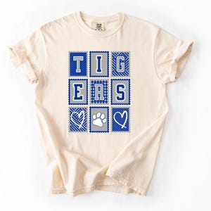 May include: A cream-colored short-sleeve t-shirt featuring a graphic print of nine square postage stamp-style frames. The frames contain the letters T, I, G, E, R, S, two white heart outlines, and a white paw print in blue, gray, and white.