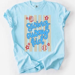 May include: Light blue t-shirt with the text "Swing Away Kid" in a playful, blue font. The design includes a baseball, glove, and stars in red. The shirt has short sleeves and a crew neck.
