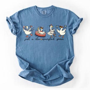 May include: A blue t-shirt featuring a graphic of four white geese in various poses with patriotic accessories. The text below the geese reads "just a star-spangled goose" with a small orange star icon replacing the hyphen in the word star-spangled.