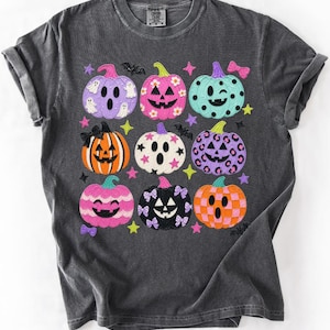 May include: A charcoal gray t-shirt featuring a grid of nine colorful jack-o'-lanterns. The pumpkins display various patterns including ghosts, flowers, leopard print, and checkers in shades of pink, orange, purple, teal, and black, accented by stars.