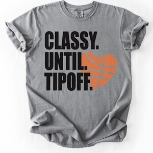 May include: A gray t-shirt with the words "CLASSY. UNTIL. TIPOFF." in black block letters. An orange basketball graphic is on the right side of the text. The shirt has a crew neck and short sleeves.