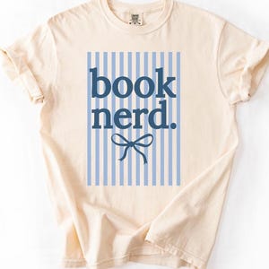 May include: Cream-colored t-shirt with the words "book nerd." in navy blue, set against a light blue and white striped background. A navy blue bow is below the text. The shirt has short sleeves and a crew neck.