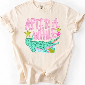 May include: A cream-colored short-sleeve t-shirt featuring a graphic of a green alligator and the pink text "AFTER A WHILE" with yellow and pink star accents. The design includes a small blue water patch and a pink flower beneath the alligator.