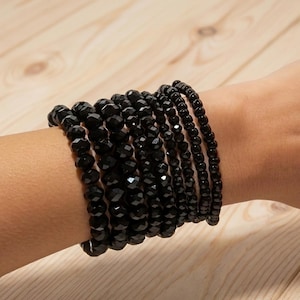 Pode incluir: Uma coleção de pulseiras de contas pretas de vários tamanhos e estilos. As pulseiras são feitas de contas facetadas e pequenas contas redondas. As pulseiras são empilhadas em um pulso.