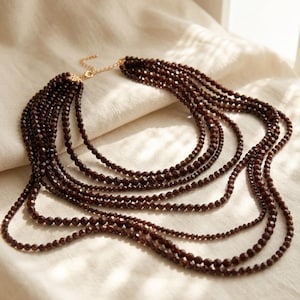 May include: A multi-strand necklace featuring small, faceted, dark brown beads. The necklace has a gold-colored clasp and is displayed on a cream-colored fabric. The strands cascade in a layered effect, creating a statement piece.