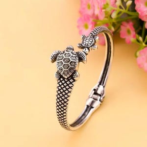 May include: A silver-toned hinged bangle bracelet featuring two detailed turtle charms. The bracelet has a textured, scale-like design on the turtle's base and a clasp closure. The background is a soft yellow with pink flowers.