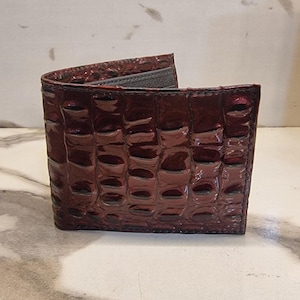 May include: A burgundy bi-fold wallet with a glossy, textured crocodile pattern. The wallet is open, revealing a grey interior and card slots. The leather has a rich, dark red hue.
