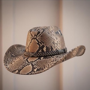 May include: Cowboy hat with a snakeskin pattern in brown and beige tones. The hat has a braided band with a silver-coloured buckle. The brim is curved, and the crown is tall, typical of a Western-style hat.