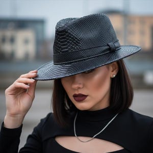 Op de afbeelding: Een zwarte fedora hoed met een textuur die op slangenleer lijkt. De hoed heeft een gestructureerde kroon en een band. De rand is breed en licht omhoog gebogen. Een stijlvol modeaccessoire.