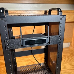 Puede incluir: Un gabinete de rack para servidores de metal negro con un soporte de montaje horizontal que sostiene un pequeño dispositivo electrónico gris rectangular con una pantalla oscura y dos antenas verticales, conectado por un cable de alimentación negro.