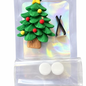 May include: A green Christmas tree air freshener clip featuring a yellow star topper, red and yellow ornaments, and a brown trunk. The set includes a black plastic vent clip and two white circular scent pads, all packaged in a clear iridescent bag.
