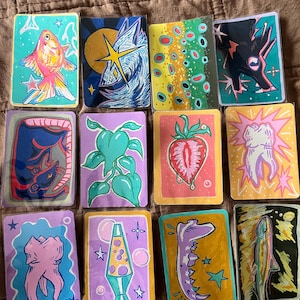 Może przedstawiać: Kolekcja dwunastu ręcznie malowanych kart artystycznych. Każda karta przedstawia unikalny wzór, w tym rybę, wilka i truskawkę. Prostokątne karty mają malowane obramowania i są ułożone na brązowej powierzchni.