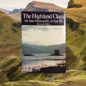 May include: A book titled "The Highland Clans" by Sir Iain Moncreiffe of that Ilk, Revised Edition. The cover features a scenic landscape with a loch, a castle, and mountains. The background shows rolling green hills and a winding road.