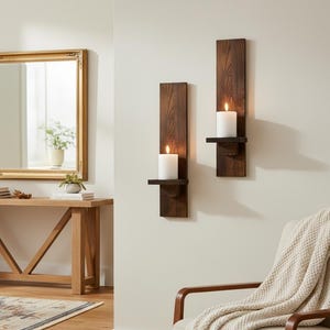 Puede incluir: Dos apliques de pared de madera marrón oscuro, cada uno con una vela blanca encendida. Los apliques están montados en una pared de color claro. Los apliques tienen un estilo rústico y de granja. Las velas son cilíndricas y proporcionan un brillo cálido.