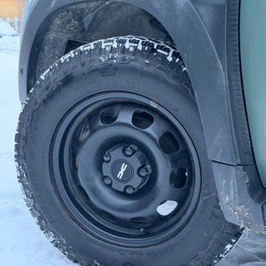 May include: Close-up of a black car wheel with a tire. The wheel is mounted on a green vehicle. The tire has a rugged tread pattern, and the wheel has a black hubcap with a white logo. Snow is visible on the tire and ground.