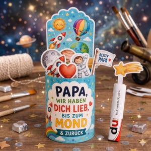 Puede incluir: Un recipiente cilíndrico azul decorado con ilustraciones espaciales que incluyen cohetes, planetas y un sol. El texto dice: "PAPA, WIR HABEN DICH LIEB, BIS ZUM MOND & ZURÜCK." Una pequeña bandera blanca adjunta muestra "BESTER PAPA" con un corazón.
