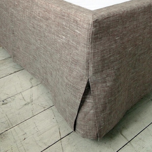 Könnte beinhalten: Ein Bett mit weißem Laken und einem braunen Bettrock aus Leinen. Der Bettrock hat ein maßgeschneidertes Design mit einer geteilten Ecke. Der Raum hat eine neutrale Farbpalette mit einer hellblauen Wand und einem Holzregal.