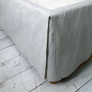 以下が含まれることがあります： 仕立てられたデザインのライトグレーのベッドスカート。ベッドスカートは柔らかく、質感のある生地で作られており、コーナーが分かれたデザインが特徴です。ベッドは白いシーツで覆われています。ベッドは、水色の壁のある部屋にあります。