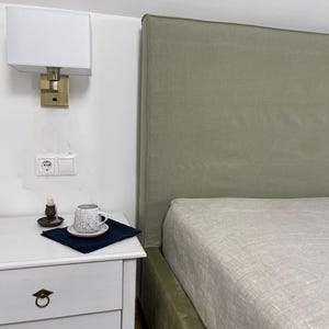 Könnte beinhalten: Ein Schlafzimmer mit einem weißen Nachttisch, einer Lampe und einem Bett mit einem salbeigrünen Kopfteil. Eine Tasse und Untertasse mit Sprenkeln liegen auf einer dunkelblauen Serviette. Das Bett hat ein helles Leinenlaken.