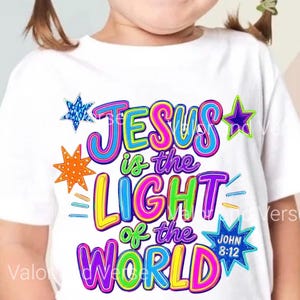 May include: A white short-sleeved t-shirt featuring a colorful graphic print with the text "JESUS is the LIGHT of the WORLD" and "JOHN 8:12". The design includes multi-colored lettering in pink, blue, yellow, and green, accented by various star shapes.