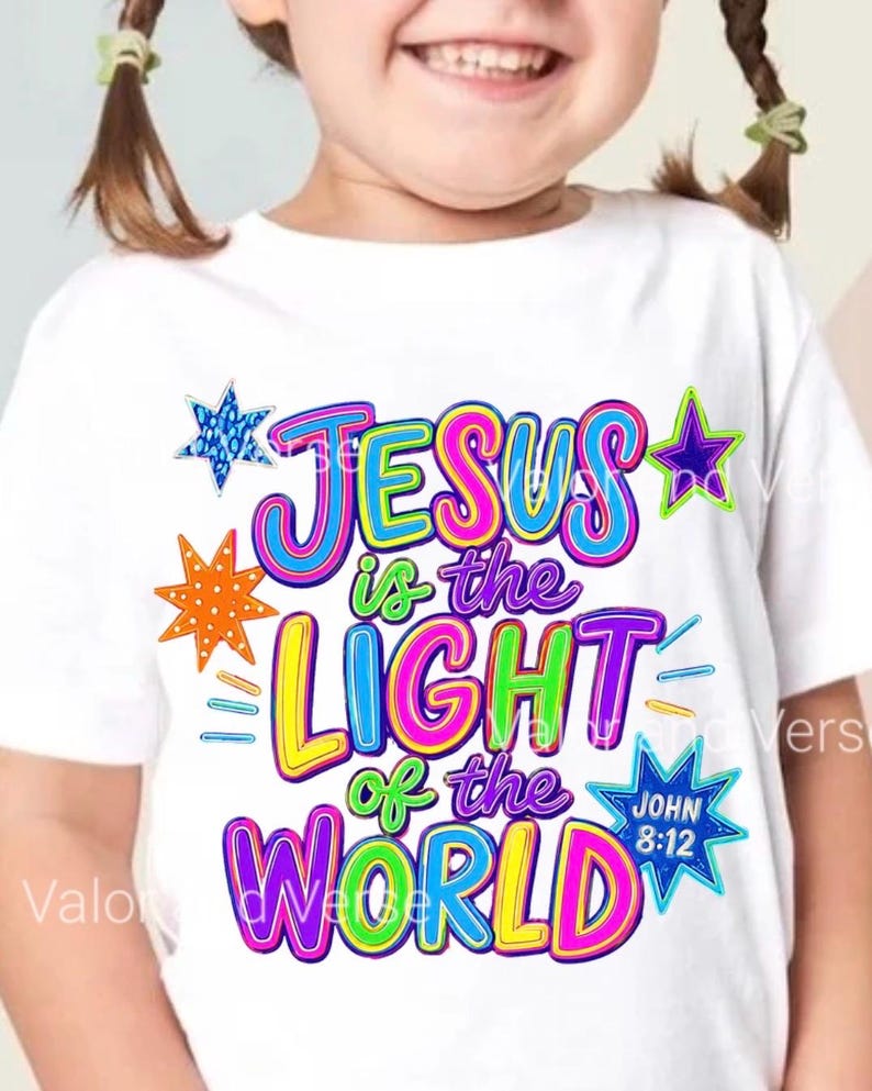 May include: A white short-sleeved t-shirt featuring a colorful graphic print with the text "JESUS is the LIGHT of the WORLD" and "JOHN 8:12". The design includes multi-colored lettering in pink, blue, yellow, and green, accented by various star shapes.