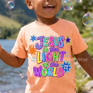 May include: A peach-colored short-sleeve t-shirt featuring a colorful graphic with the text 'JESUS is the LIGHT of the WORLD' and 'JOHN 8:12'. The design includes various star shapes in blue, orange, and purple, with bright neon-style lettering.