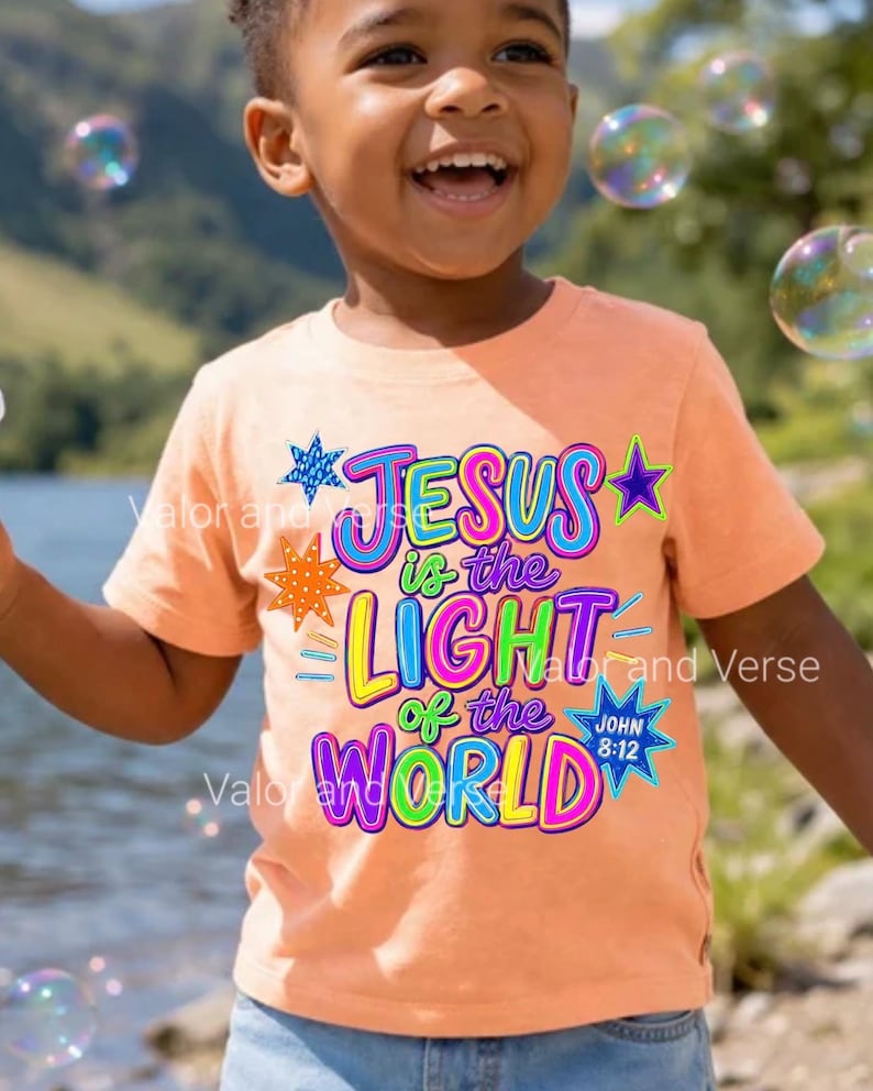 May include: A peach-colored short-sleeve t-shirt featuring a colorful graphic with the text 'JESUS is the LIGHT of the WORLD' and 'JOHN 8:12'. The design includes various star shapes in blue, orange, and purple, with bright neon-style lettering.