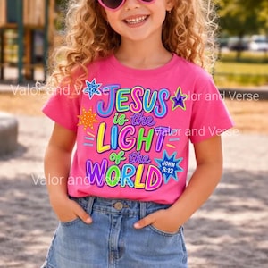 May include: A bright pink short-sleeve t-shirt featuring a colorful graphic print that reads "JESUS is the LIGHT of the WORLD" with "JOHN 8:12" in a blue star. The design includes various multi-colored stars and decorative accents.