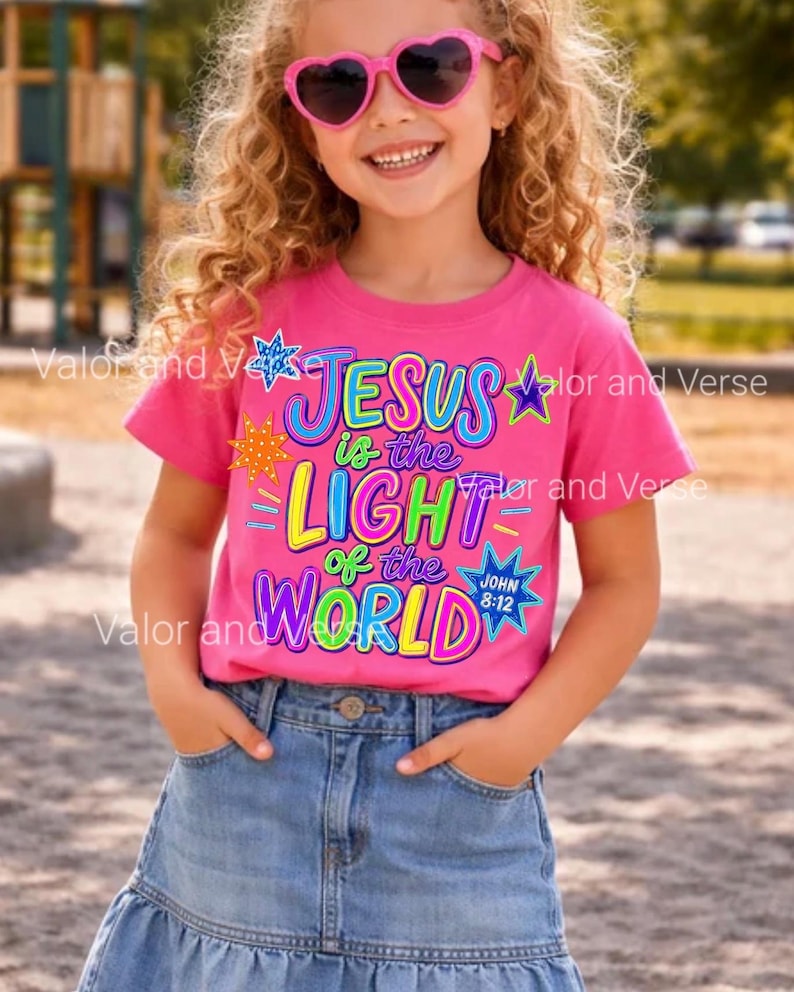 May include: A bright pink short-sleeve t-shirt featuring a colorful graphic print that reads "JESUS is the LIGHT of the WORLD" with "JOHN 8:12" in a blue star. The design includes various multi-colored stars and decorative accents.