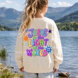 May include: A cream-colored crewneck sweatshirt featuring a vibrant, multi-colored graphic on the back that reads "JESUS is the LIGHT of the WORLD" with "JOHN 8:12" and various star-shaped accents in bright pink, blue, yellow, and purple.