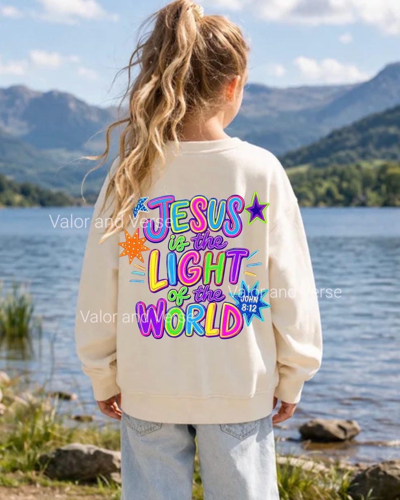 May include: A cream-colored crewneck sweatshirt featuring a vibrant, multi-colored graphic on the back that reads "JESUS is the LIGHT of the WORLD" with "JOHN 8:12" and various star-shaped accents in bright pink, blue, yellow, and purple.