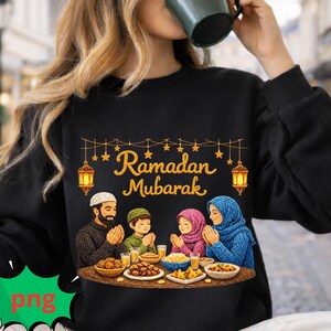 May include: Black sweatshirt featuring a family celebrating Ramadan. The design includes the text "Ramadan Mubarak", lanterns, and a family praying at a table with food. The family members are wearing traditional head coverings. The image is colorful.