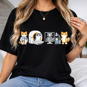 May include: Black t-shirt featuring a graphic of cartoon cats in medical settings. The design includes a cat doctor, a cat in an MRI machine, a cat's x-ray, and a cat technician. The cats are orange, white, and gray.