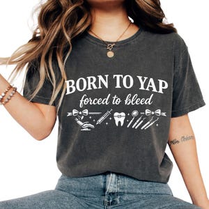 以下が含まれることがあります： 「BORN TO YAP forced to bleed」の白文字がプリントされた濃いグレーのTシャツ。デザインには、歯科をテーマにしたグラフィックが含まれています。クルーネックの柔らかい素材のTシャツです。